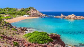 A coastal landscape with a beach, clear blue waters, rocky outcrops, and a hill with greenery. - Aparthotel Sol Parc (Mercadal)