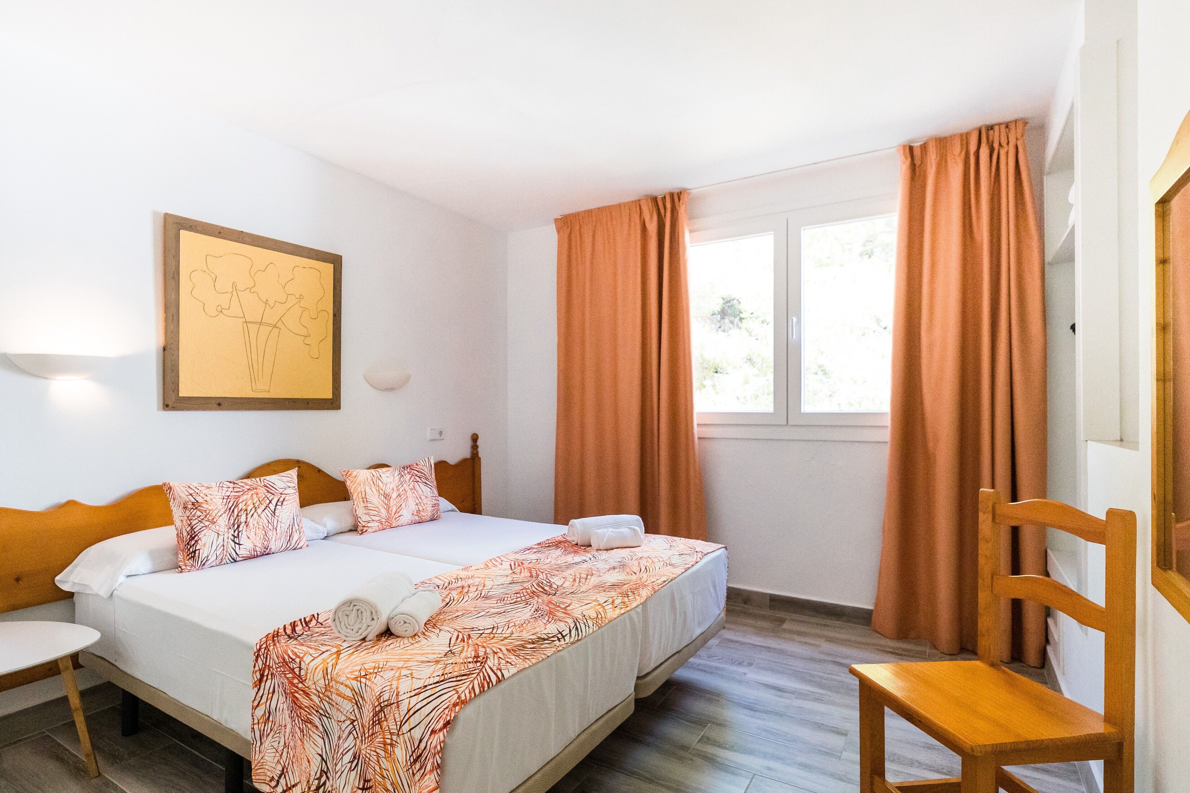 A double bed with a floral bedspread, a wooden chair, a bedside table, and a window with curtains.