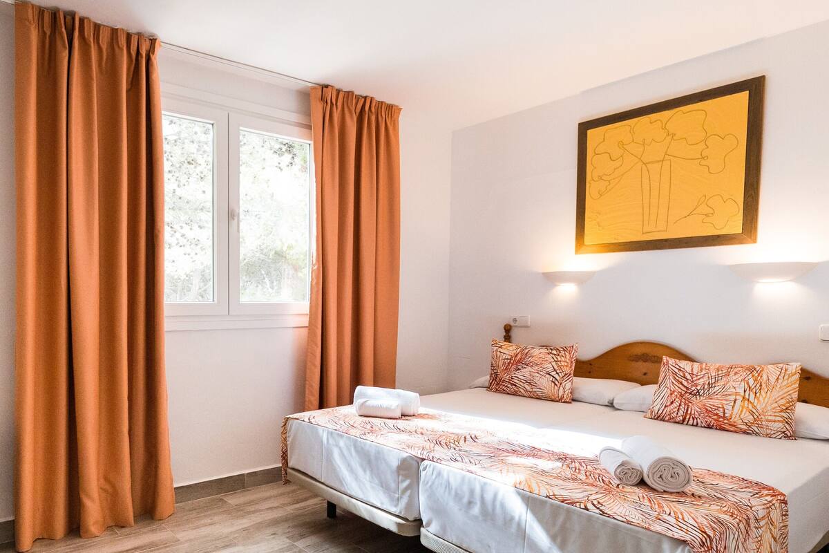 A hotel room with a bed, a chair, a window with curtains, and a wall-mounted light.