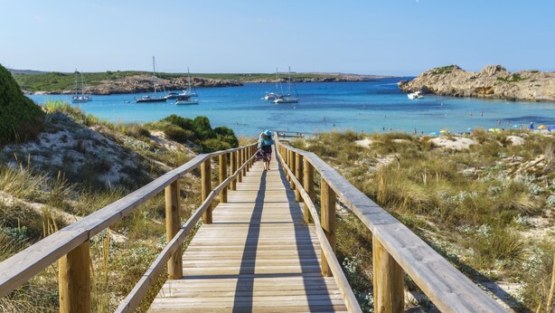 A wooden boardwalk leading to a beach with boats and clear blue water. - Aparthotel Sol Parc (Mercadal)