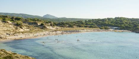 A coastal beach with clear waters, sandy shores, and a mountainous backdrop.