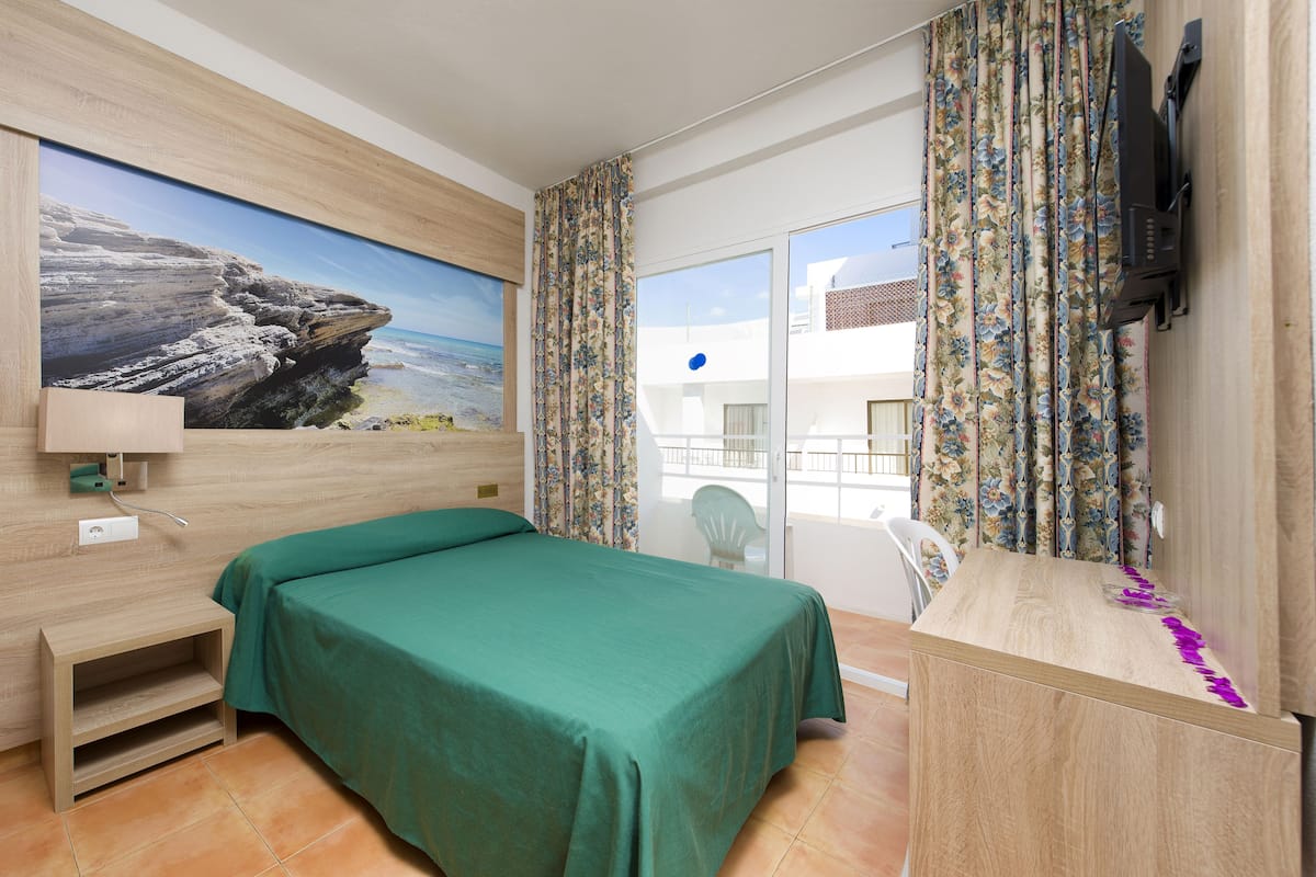 Een hotelkamer met een bed, een bureau, een televisie en een balkon met gordijnen.