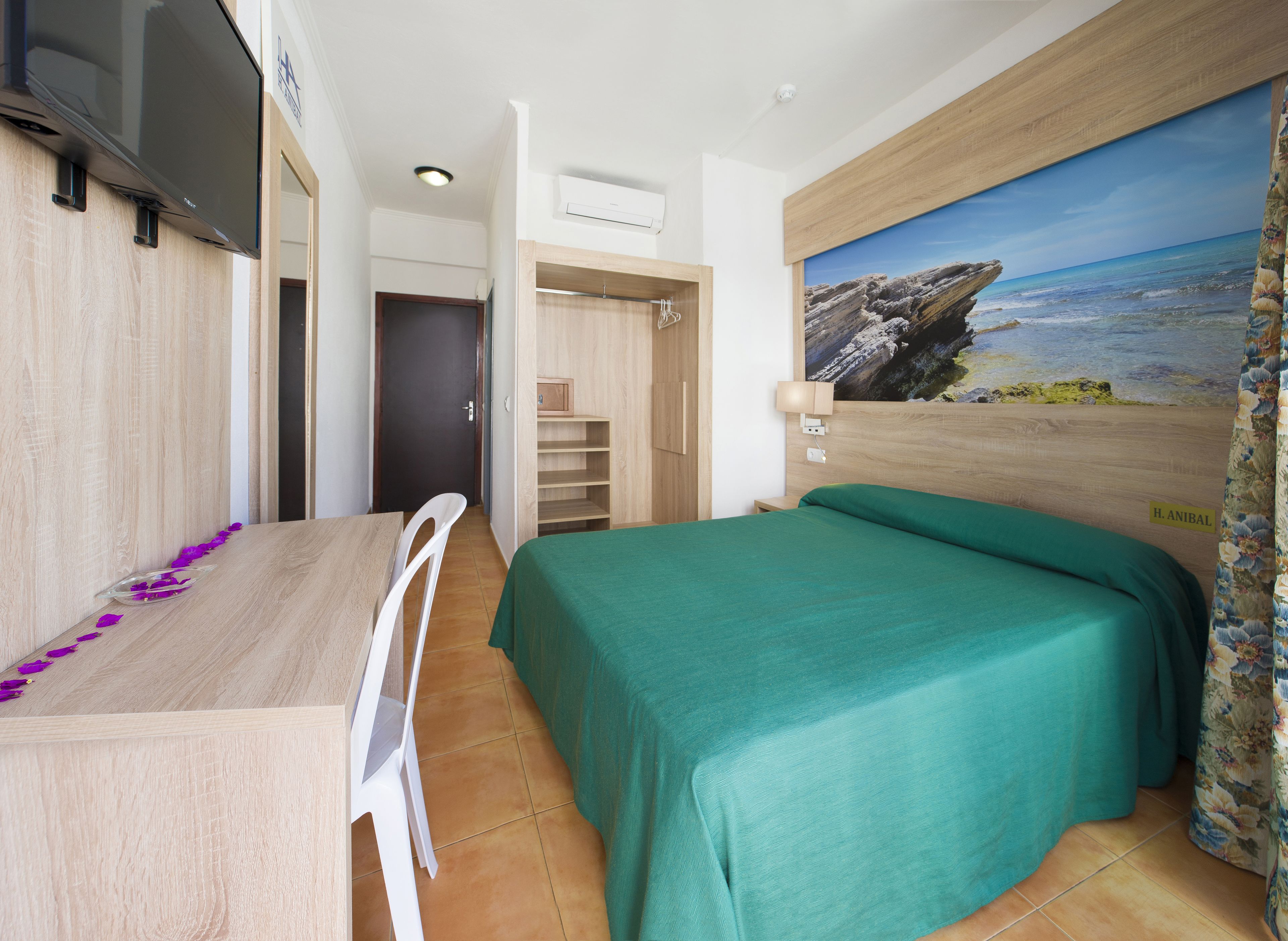 A hotel room with a bed, a desk, a chair, a TV, and a painting of a beach scene.