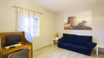 Una habitación con un escritorio de madera, una computadora, un teclado y una silla. Hay un sofá azul y una pequeña mesa blanca con una planta. En la pared, se ve una ventana con cortinas y un cuadro de una escena costera.