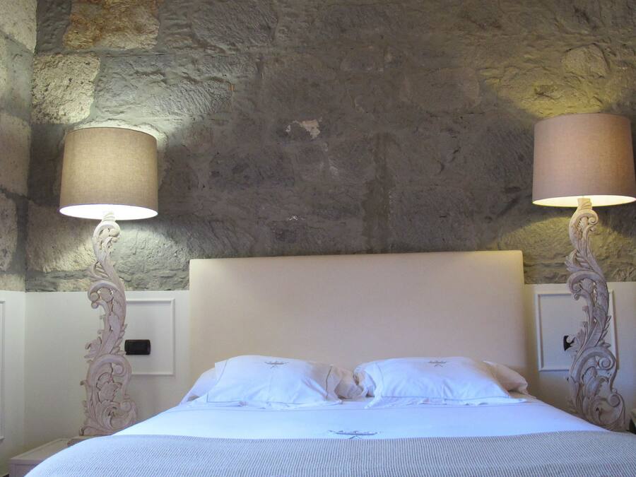 A bed with white linens, two wall-mounted lamps, and a stone wall behind.
