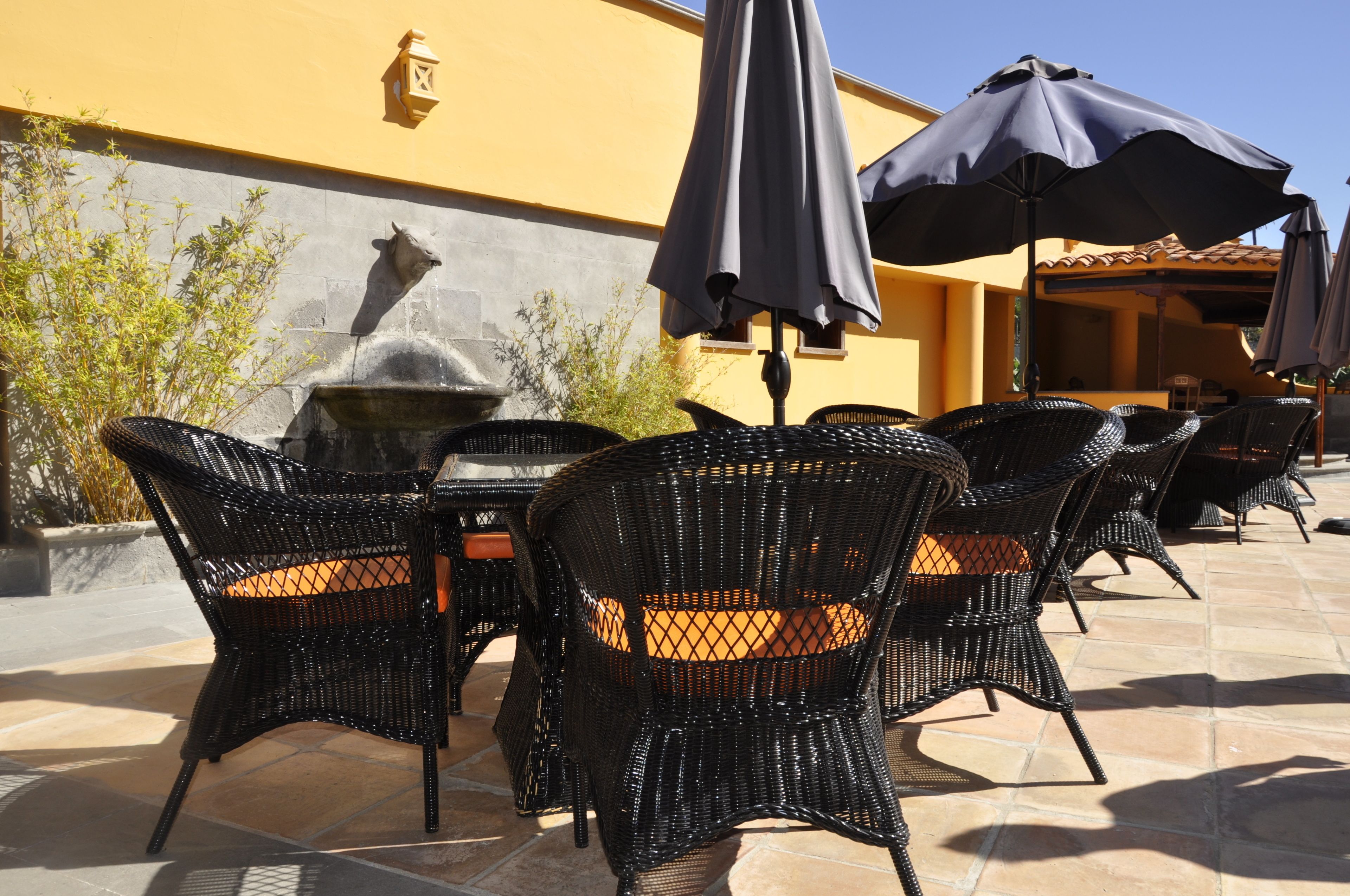 Un patio al aire libre con sillas de mimbre y sombrillas, una fuente de piedra y un edificio amarillo al fondo.
