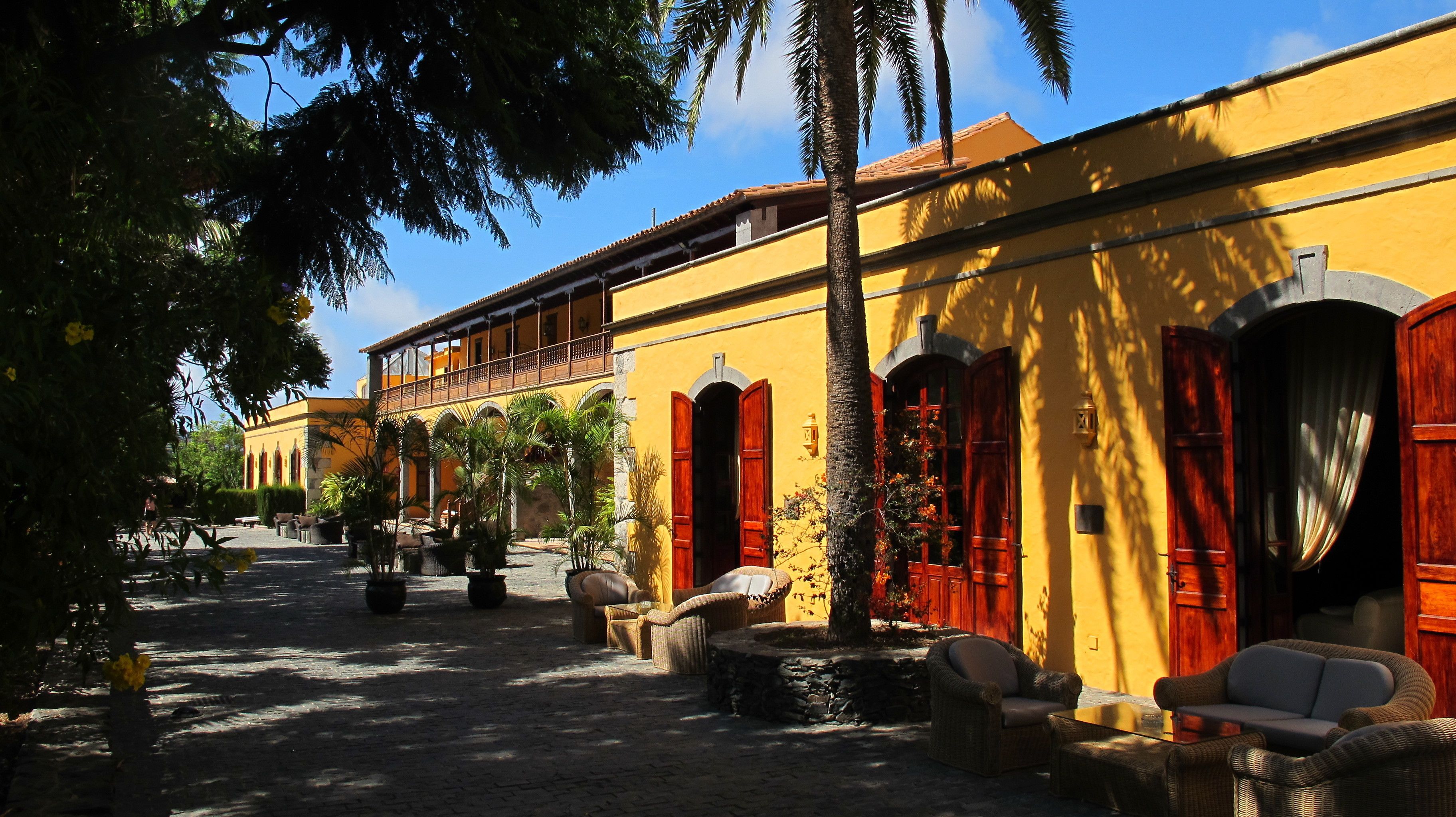 A courtyard with outdoor seating, a yellow building with wooden shutters, and a clear blue sky.