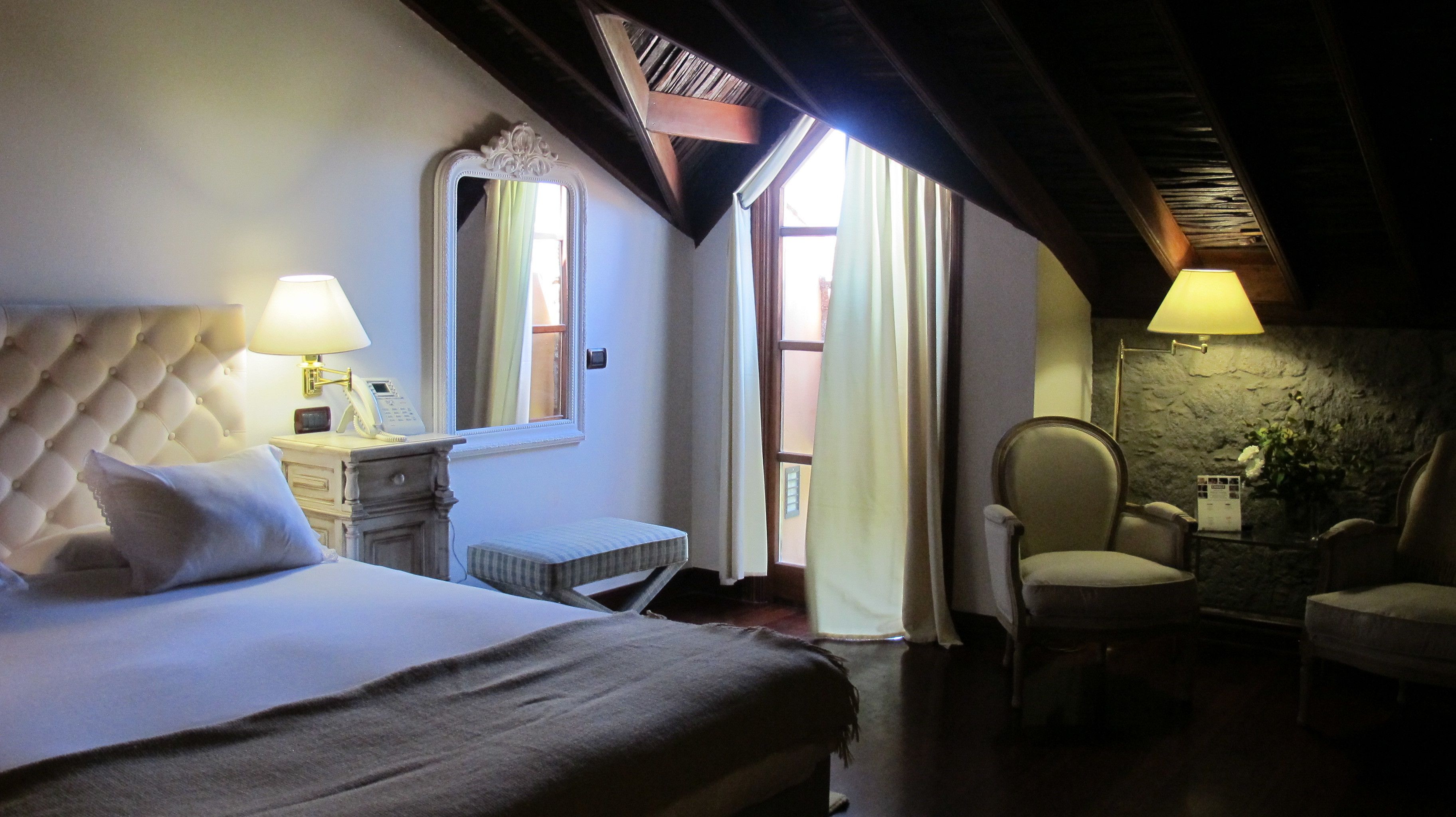 A bedroom with a large bed, a nightstand with a lamp, a chair, and a stone wall.