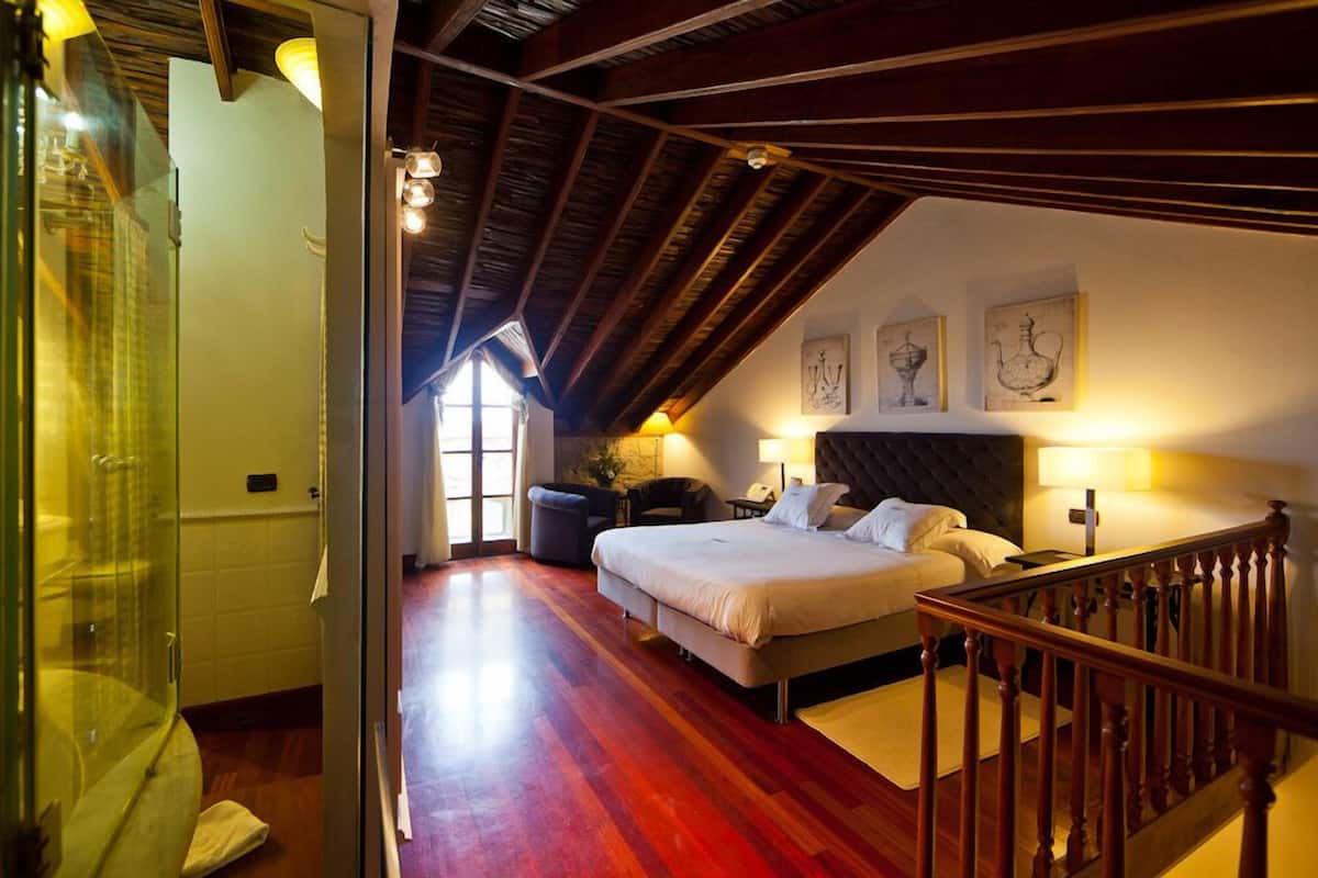 Ein Schlafzimmer mit einem großen Bett, einer Holzbalkendecke und Blick ins Freie durch ein Fenster.