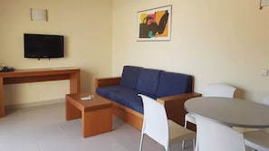 Ein Wohnzimmer mit blauem Sofa, einem Holztisch, einem Fernseher, einem Gemälde, einem weißen Sessel und einem runden Tisch.