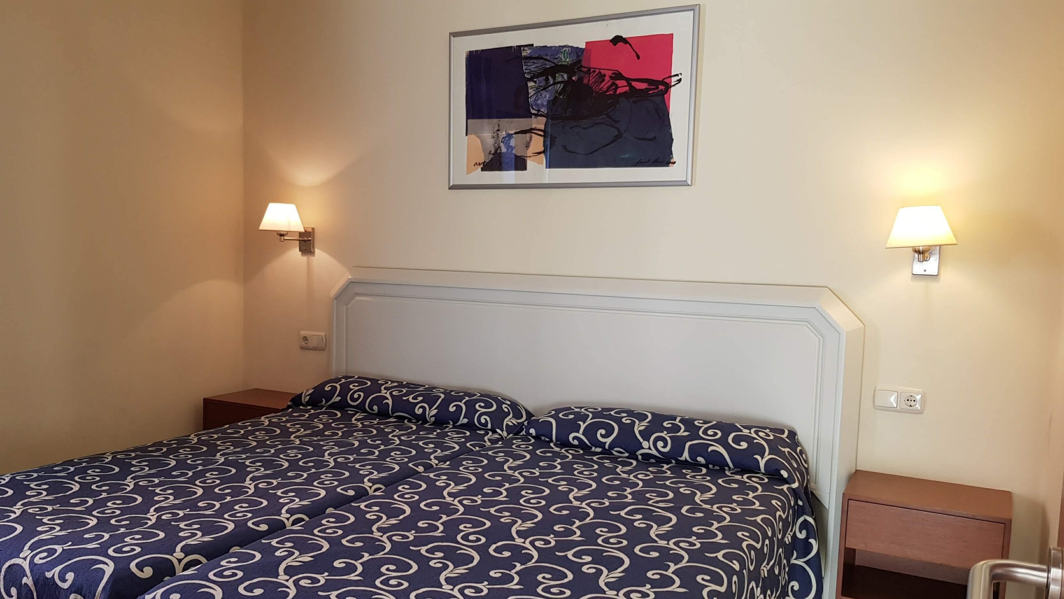 Een netjes opgemaakt bed met een geborduurde spreideken, twee bedlampen en een lijst met abstracte kunst boven het bed.