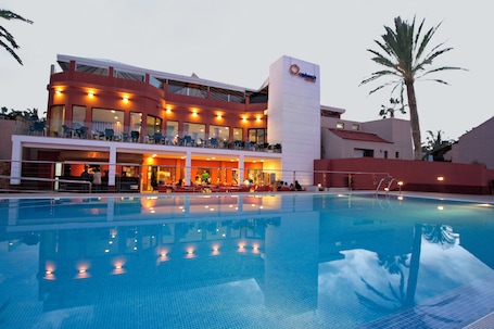 A hotel with a swimming pool, outdoor seating, and a palm tree.
