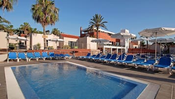 Une piscine entourée de palmiers et d’immeubles, avec des chaises longues bleues et des parasols.