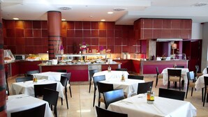 Un restaurante con varias mesas y sillas, una zona de bufé central y una cocina al fondo.