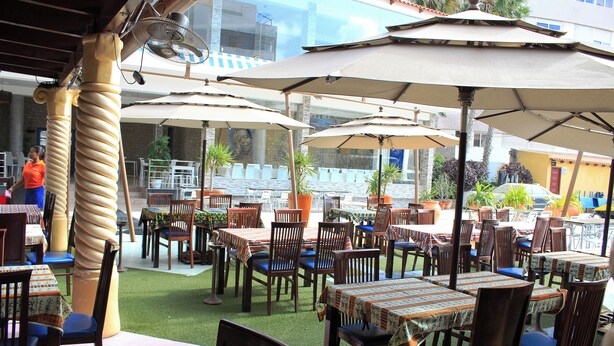 Un área de comedor al aire libre con varias mesas y sillas, cubierta por sombrillas y rodeada de plantas en macetas.