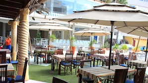 An outdoor dining area with multiple tables and chairs, covered by umbrellas, and surrounded by potted plants. - Mindel Hotel (São Vicente)