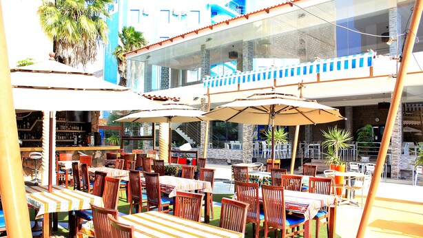 An outdoor dining area with tables and chairs, umbrellas, and a pool in the background. - Mindel Hotel (São Vicente)