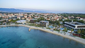 An aerial view of a coastal town with a hotel featuring a swimming pool, a beach, and a view of the mountains in the background. - Hotel Liberty (Novalja)
