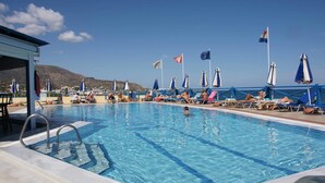 Uma piscina com espreguiçadeiras e guarda-sóis, bandeiras e montanhas ao fundo.