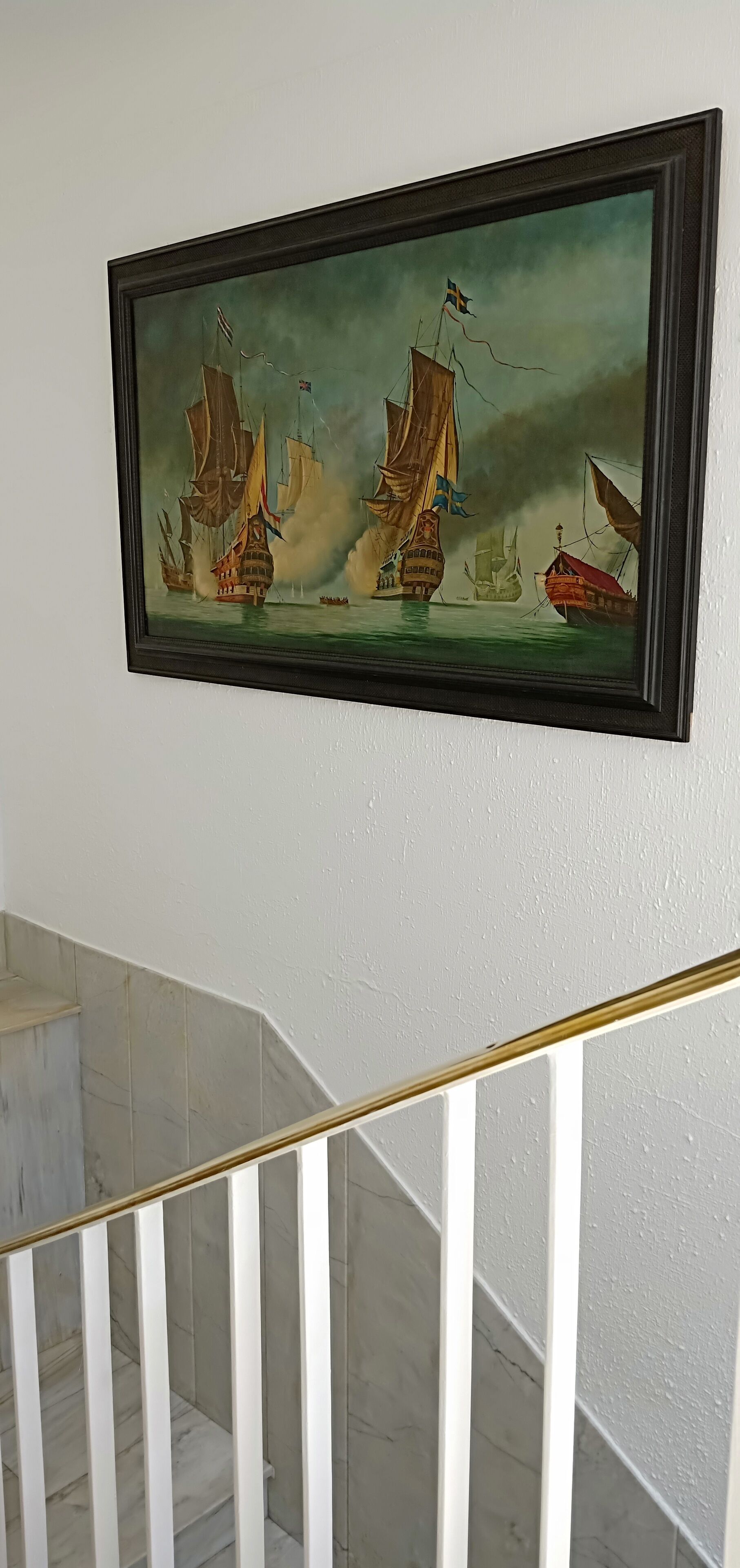 A framed maritime painting on a wall above a staircase with white railings and marble steps.