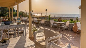 A covered outdoor seating area with tables and chairs, overlooking a beach. - Kathrin Beach Hotel (ADELIANOS KAMPOS)