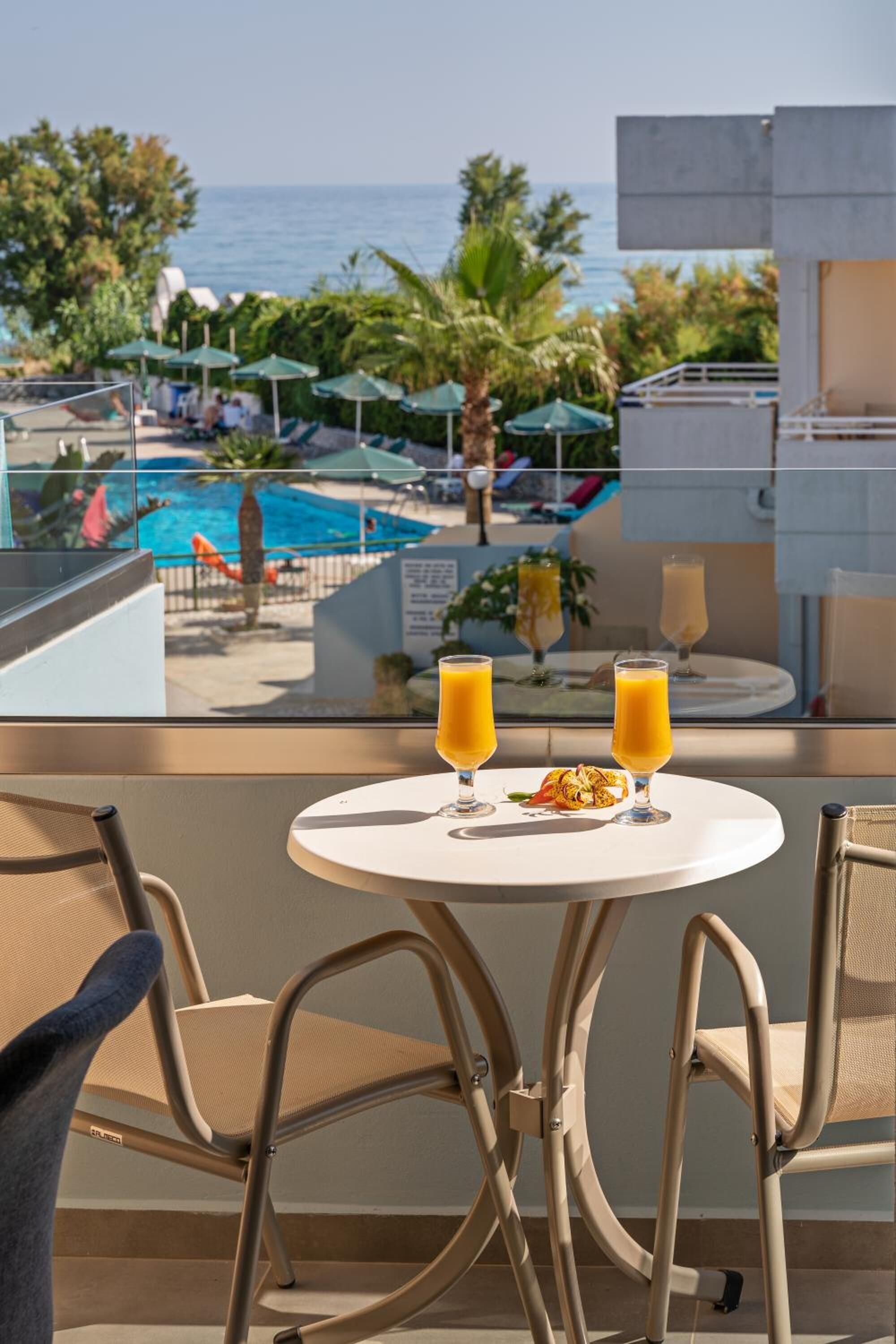 A balcony with a table, two glasses of orange juice, and a tropical view of a pool and ocean.