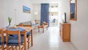 A modern living room with a sofa, dining table, and chairs, a TV on a cabinet, and a large window with curtains. - Apartamentos ES Trenc (Ses Salines)