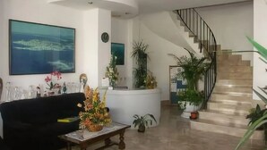 A spacious living room with a black sofa, a coffee table, and a large painting of a coastal landscape. A staircase with a black railing leads to an upper floor. - Apartamentos ES Trenc (Ses Salines)