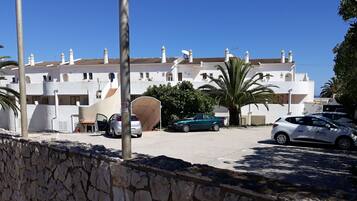 A white building with multiple balconies and satellite dishes, a stone wall in the foreground, and parked cars.