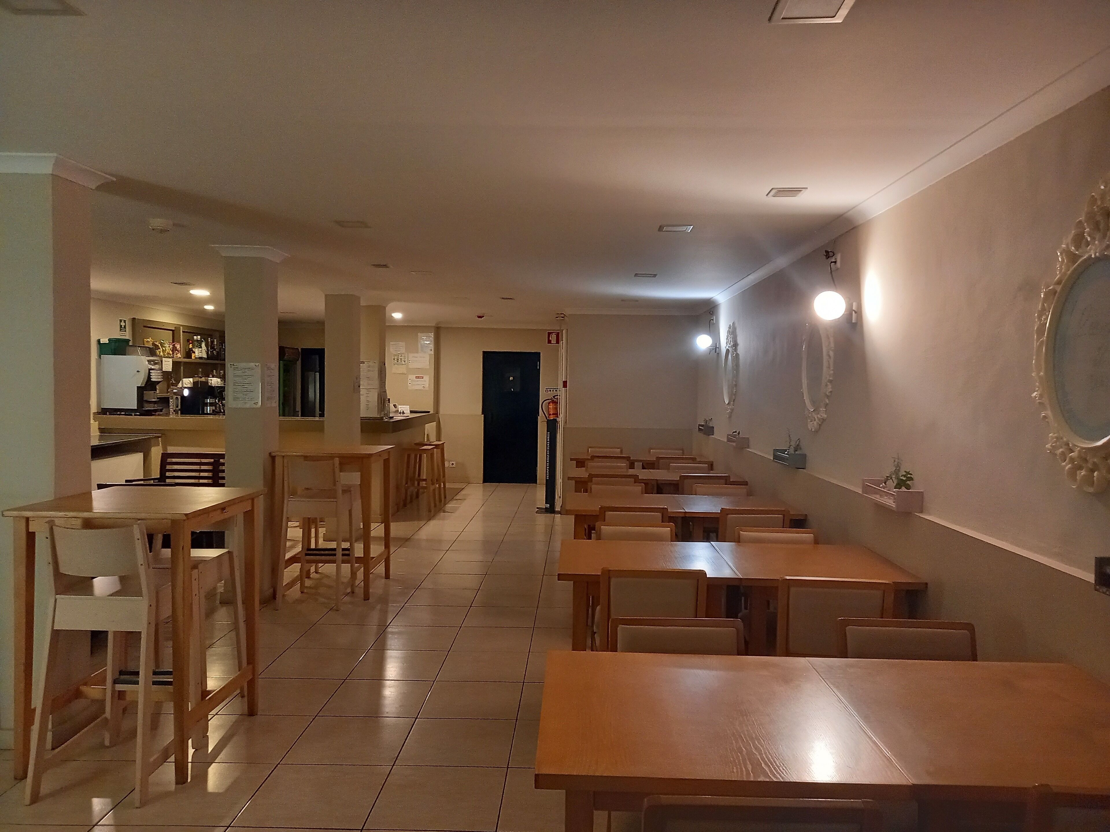 A long corridor with multiple wooden tables and chairs, leading to a kitchen area with a refrigerator and coffee machine.