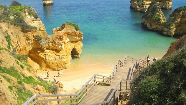 Uma área costeira com uma escadaria de madeira que desce para uma praia arenosa, rodeada por falésias rochosas e águas azuis cristalinas.