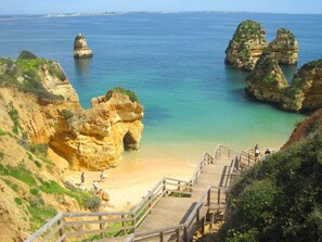 A coastal area with a wooden staircase leading down to a sandy beach, surrounded by rocky cliffs and clear blue water. - Vilamar (Lagos)
