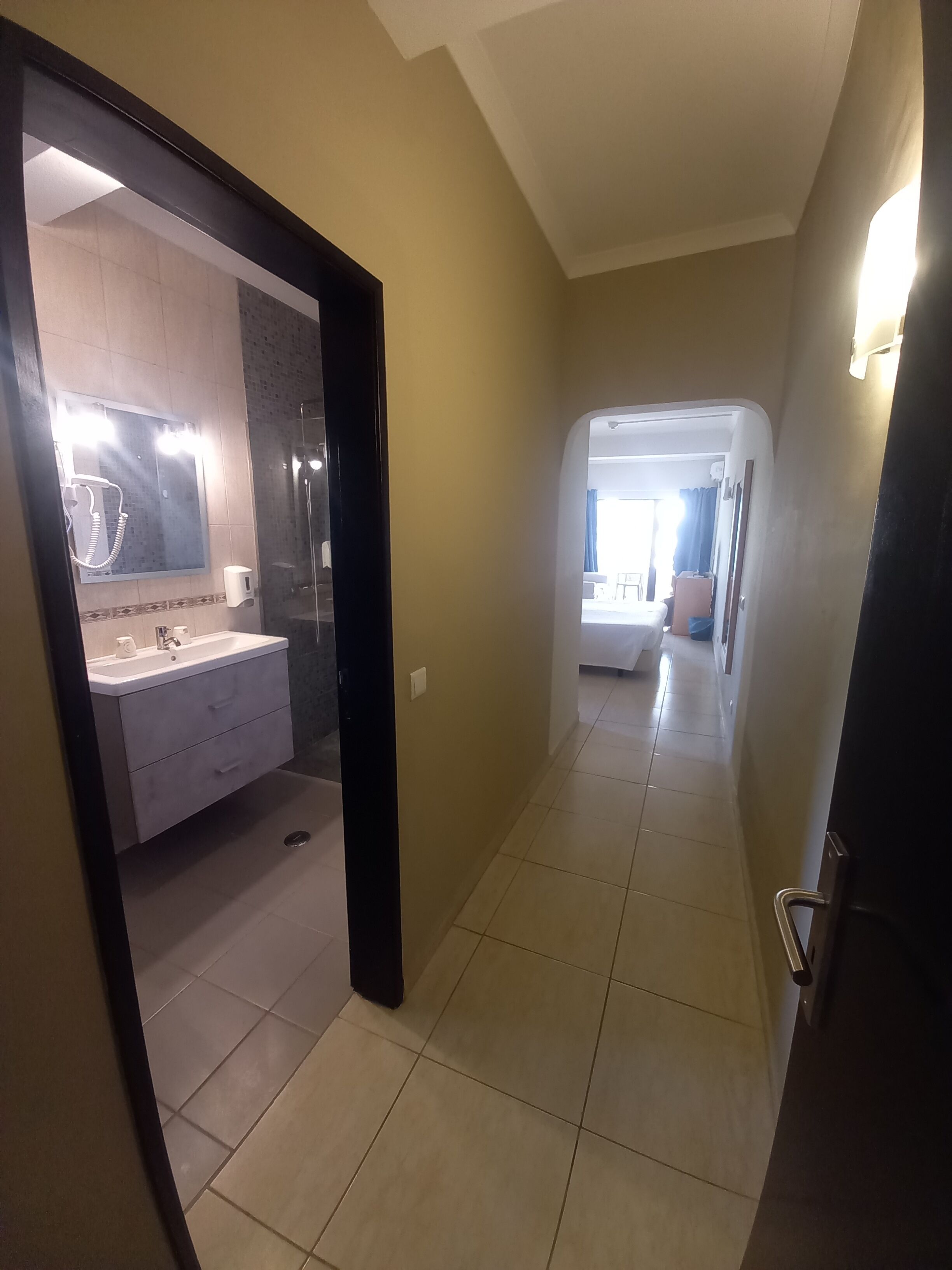 Un pasillo de hotel que lleva a un dormitorio con cama y ventana, y cuenta con un baño que incluye lavamanos, espejo y ducha.