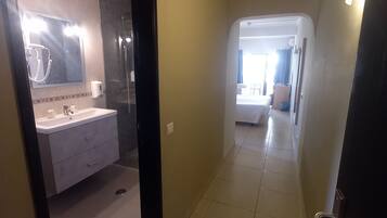 Un pasillo de hotel que lleva a un dormitorio con cama y ventana, y cuenta con un baño que incluye lavamanos, espejo y ducha.