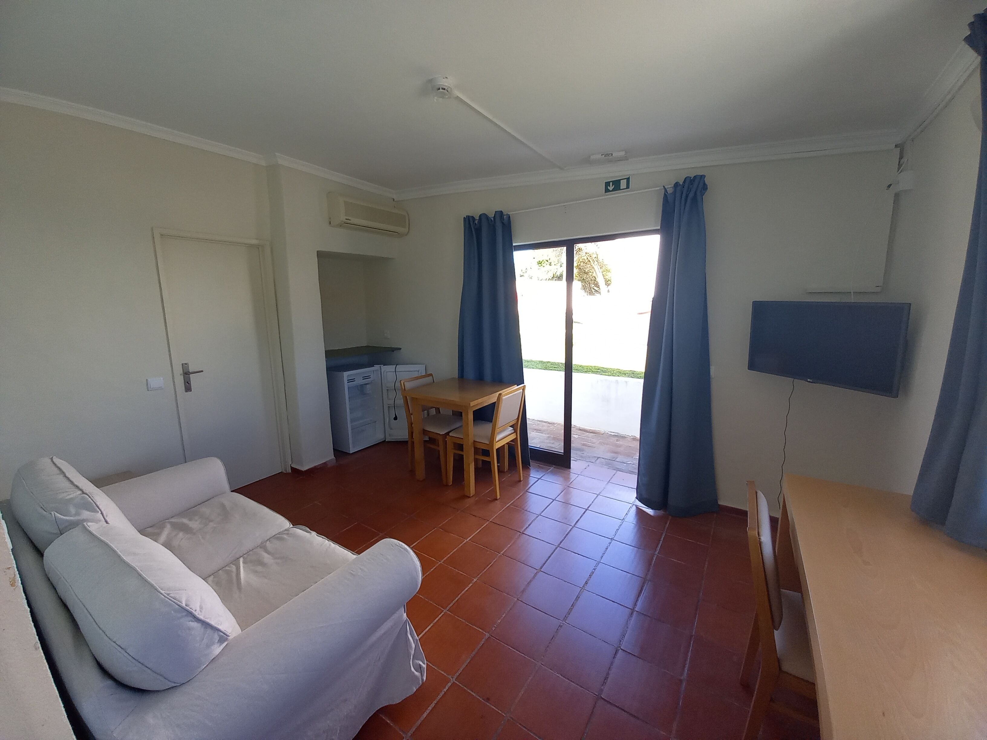 A room with a sofa, a dining table, a TV, an air conditioning unit, and a sliding glass door leading to an outdoor area.