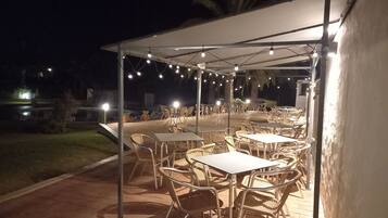 An outdoor patio area with tables and chairs under a canopy, string lights overhead, and a view of the surrounding landscape.