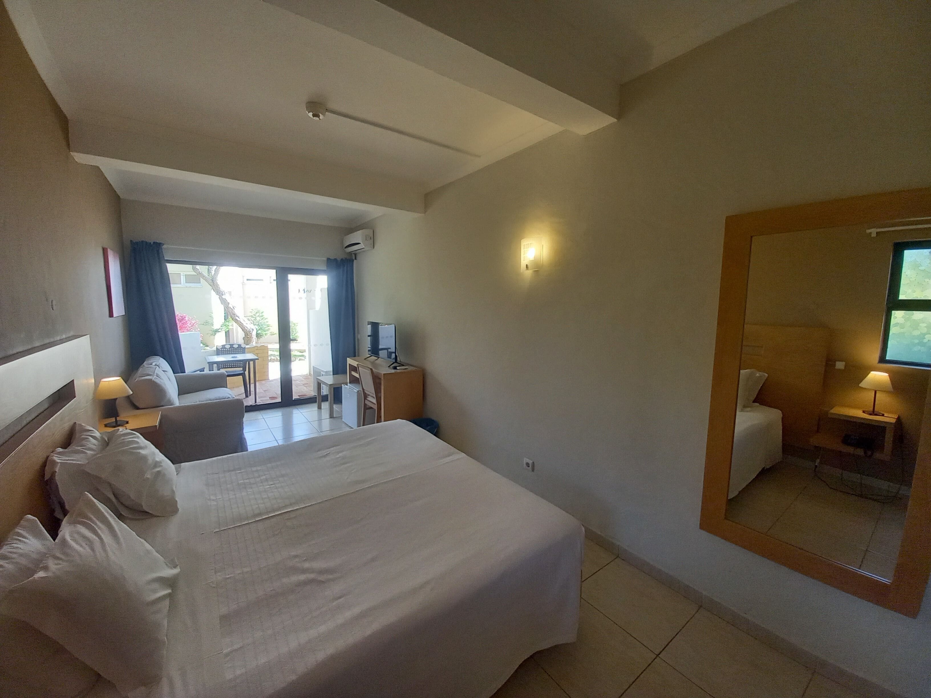 A hotel room with a large bed, a desk, a chair, a small table, and a mirror.
