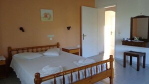 A bedroom with a wooden bed, a bedside table, a nightstand, a small wooden stool, a mirror, and a door leading to another room. - Apartments Villa Frangis (Corfu)