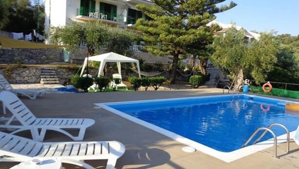 A swimming pool with lounge chairs, a white building with green shutters, and a pine tree. - Apartments Villa Frangis (Corfu)
