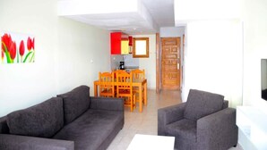 A living room with two grey sofas, a dining table with chairs, a kitchen area with red cabinets, and a wooden door. - Niza (Mogan)