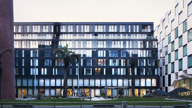 Un edificio moderno de varios pisos con fachada de vidrio, un patio central con palmeras y una entrada visible con marquesina.