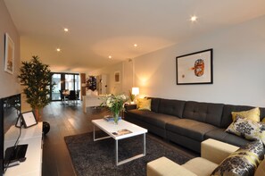 Una sala de estar moderna con un sofá, una mesa de centro y una planta grande. En el fondo, se observa un comedor con una mesa y sillas.