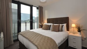 Habitación de hotel con una cama grande, una mesita de noche con lámpara y vistas al exterior a través de una puerta de cristal.