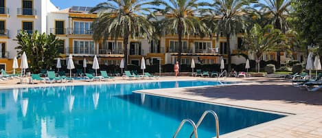 A swimming pool with lounge chairs and umbrellas, surrounded by palm trees and a hotel building.