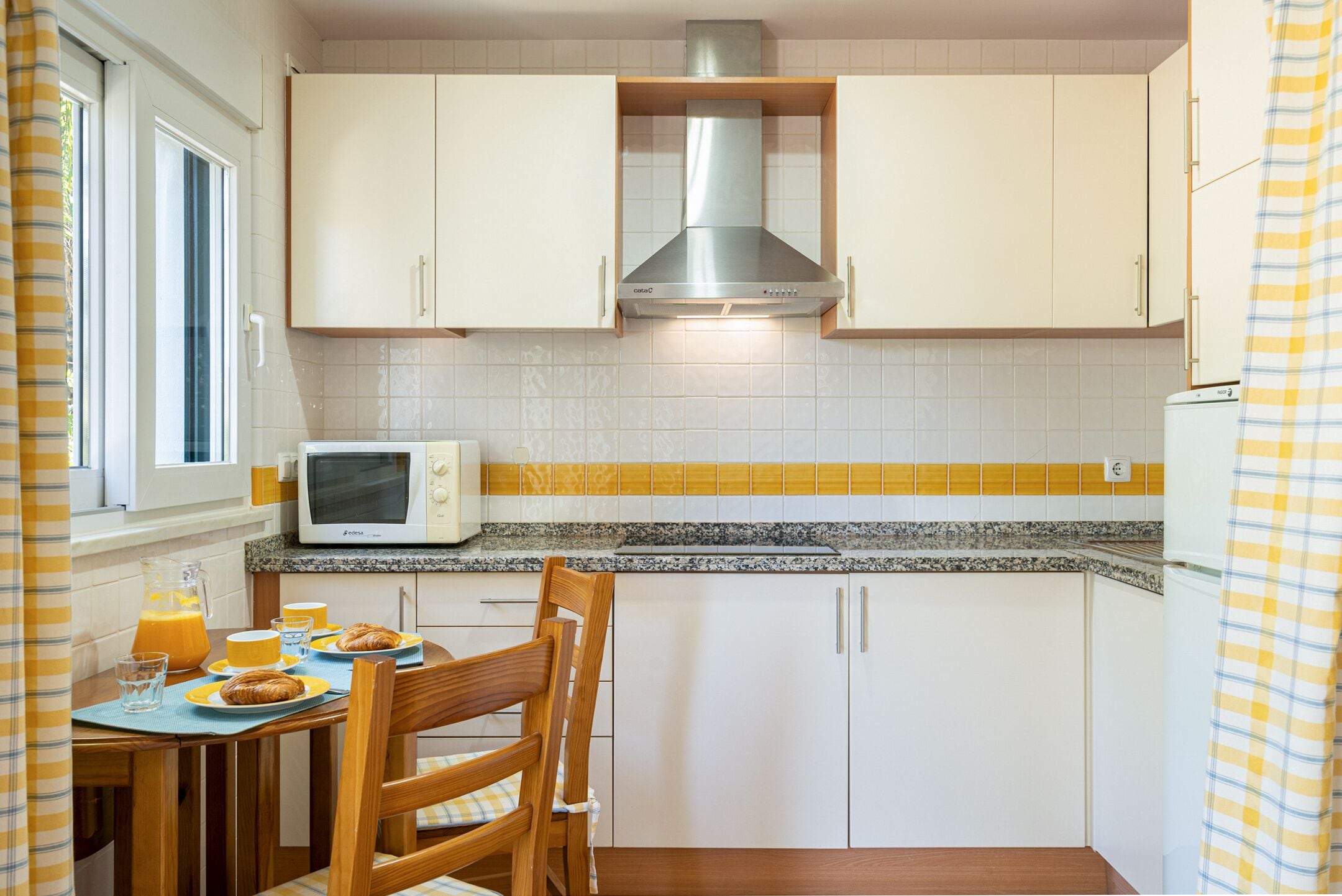 Una cucina compatta con mobili bianchi, una cappa in acciaio inossidabile, un forno a microonde e un angolo colazione con croissant e succo d'arancia.