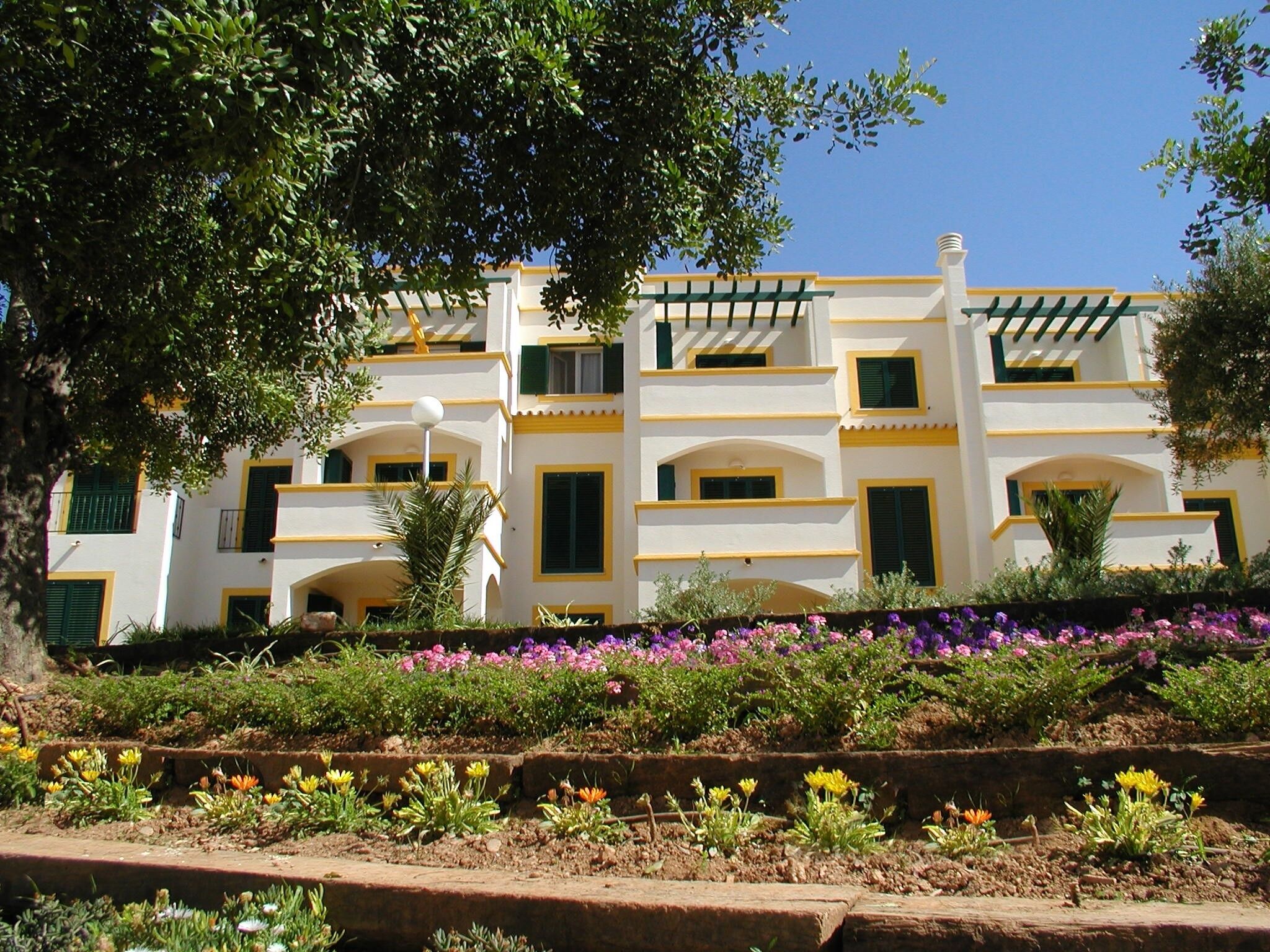 A multi-story building with balconies, a garden with various flowers, and a clear blue sky.