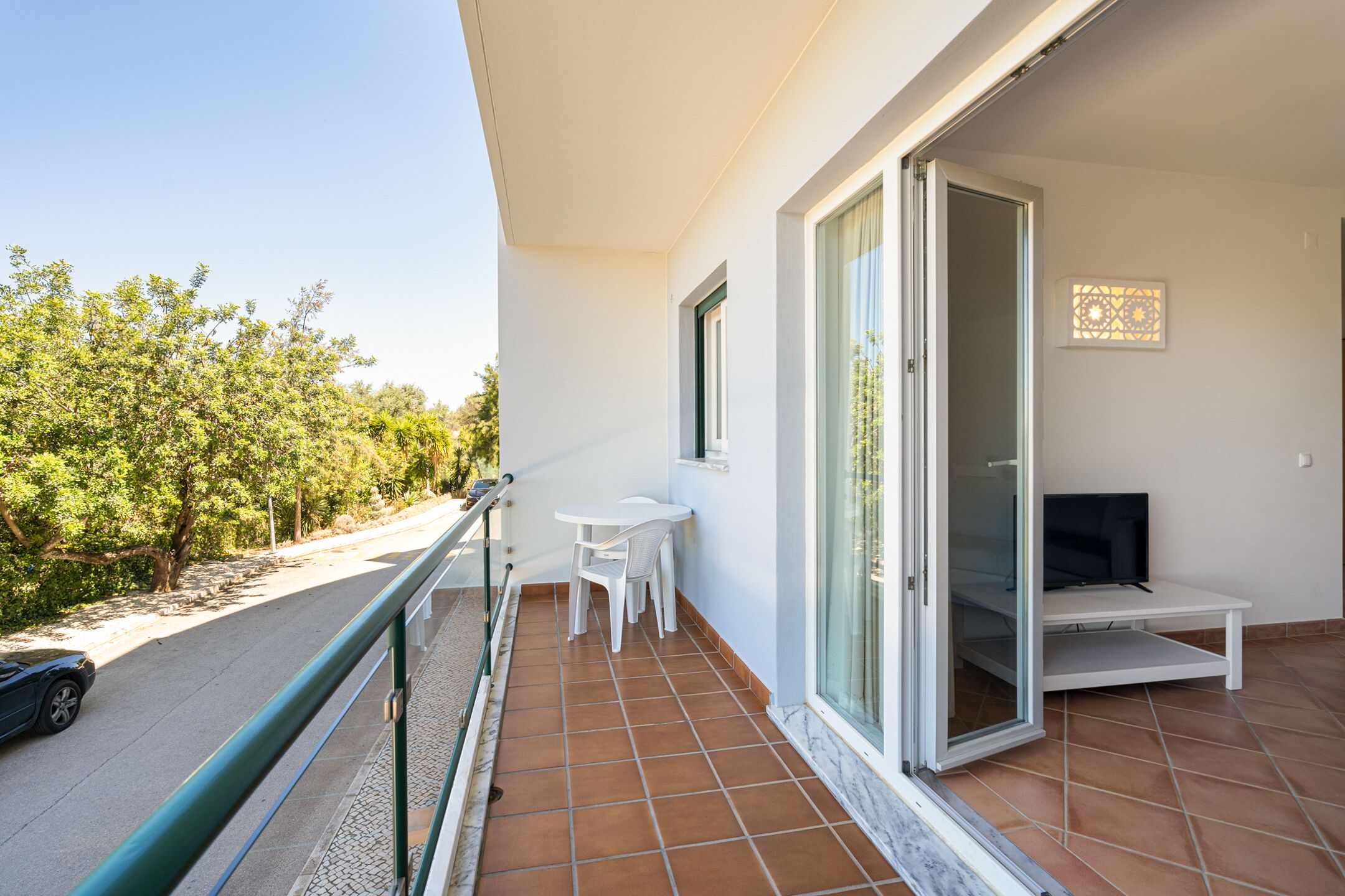 Un balcon avec une table et des chaises blanches, un téléviseur et une porte vitrée donnant sur un espace intérieur.