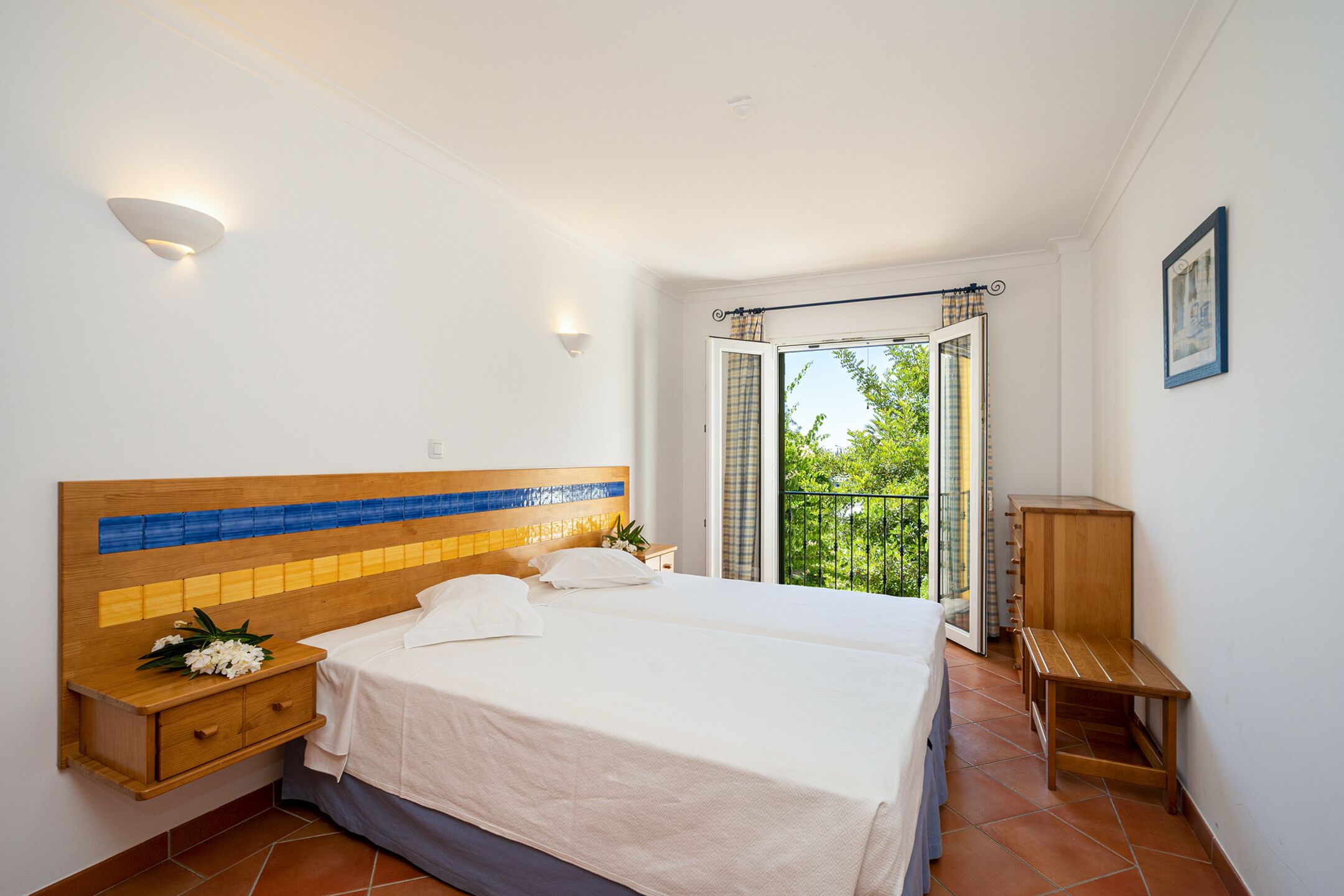 A bedroom with a large bed, a nightstand, a wooden bench, and a view of greenery through an open door.