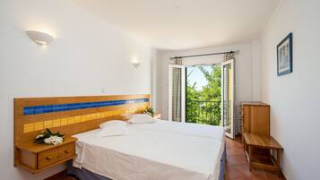 A bedroom with a large bed, a nightstand, a wooden bench, and a view of greenery through an open door.