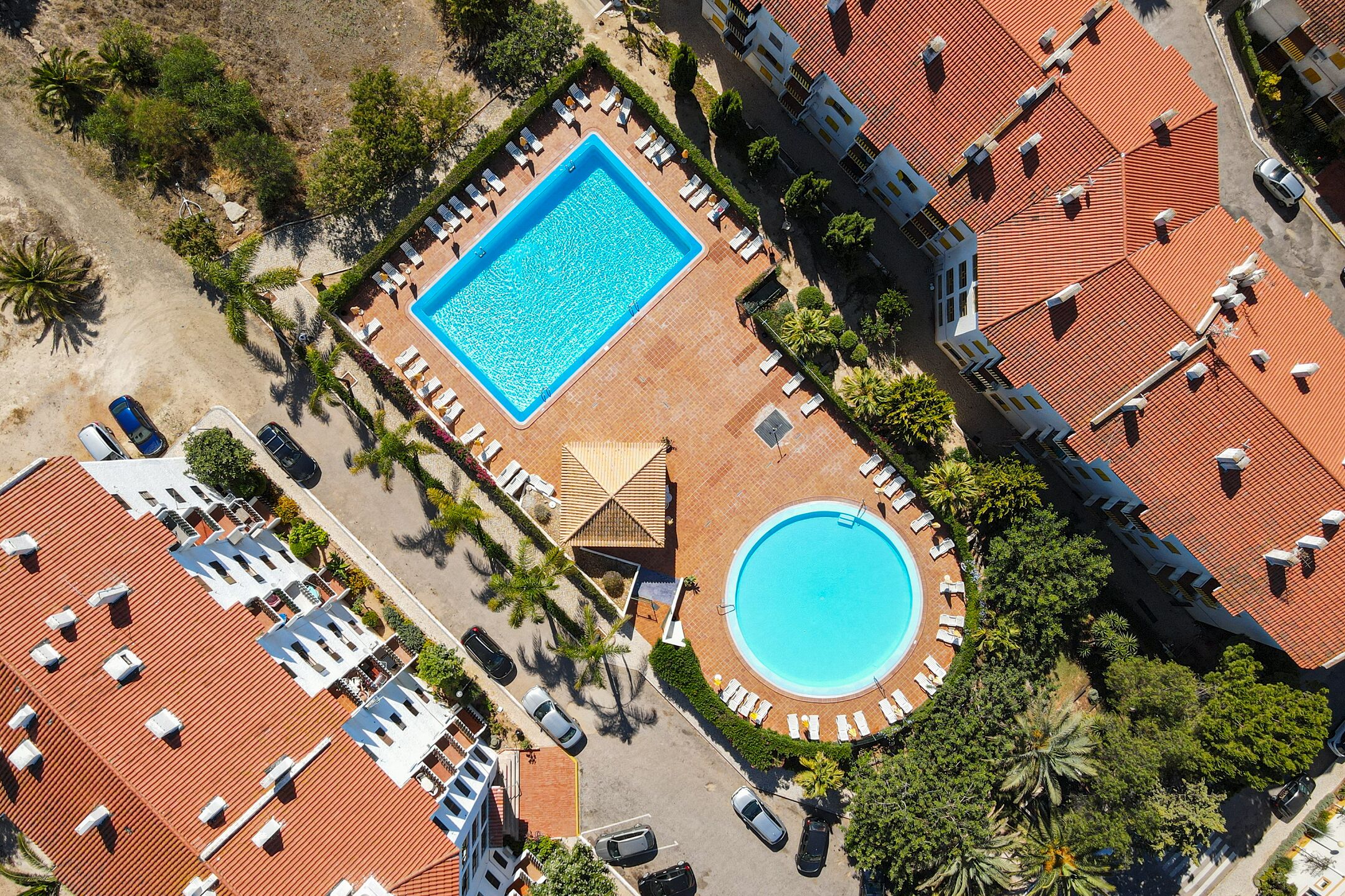 Eine Vogelperspektive zeigt ein Resort mit einem rechteckigen Swimmingpool, einem runden Pool und mehreren Liegebereichen mit Sonnenliegen.